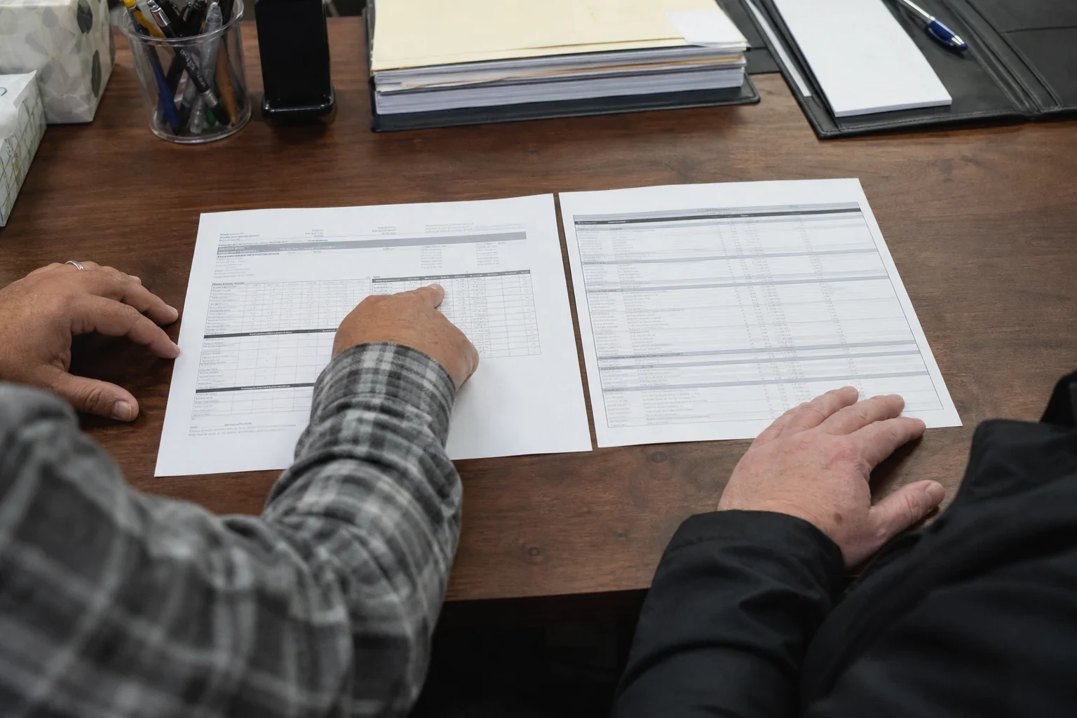 Two people at a wooden office desk, one in a plaid shirt pointing at a printed statement laid beside a coverage table, with folders and a pen cup nearby.