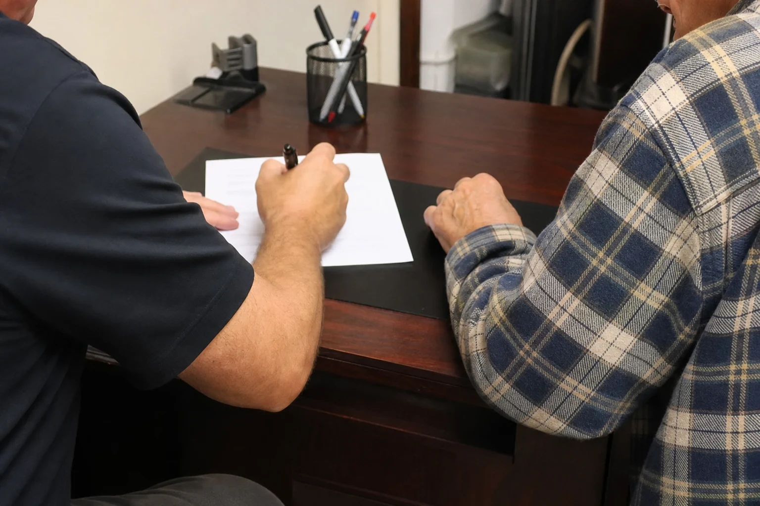 Two men seated at a wooden office desk, one in a navy shirt holding a pen over a single-page form while an older man in a plaid flannel shirt rests his hand nearby