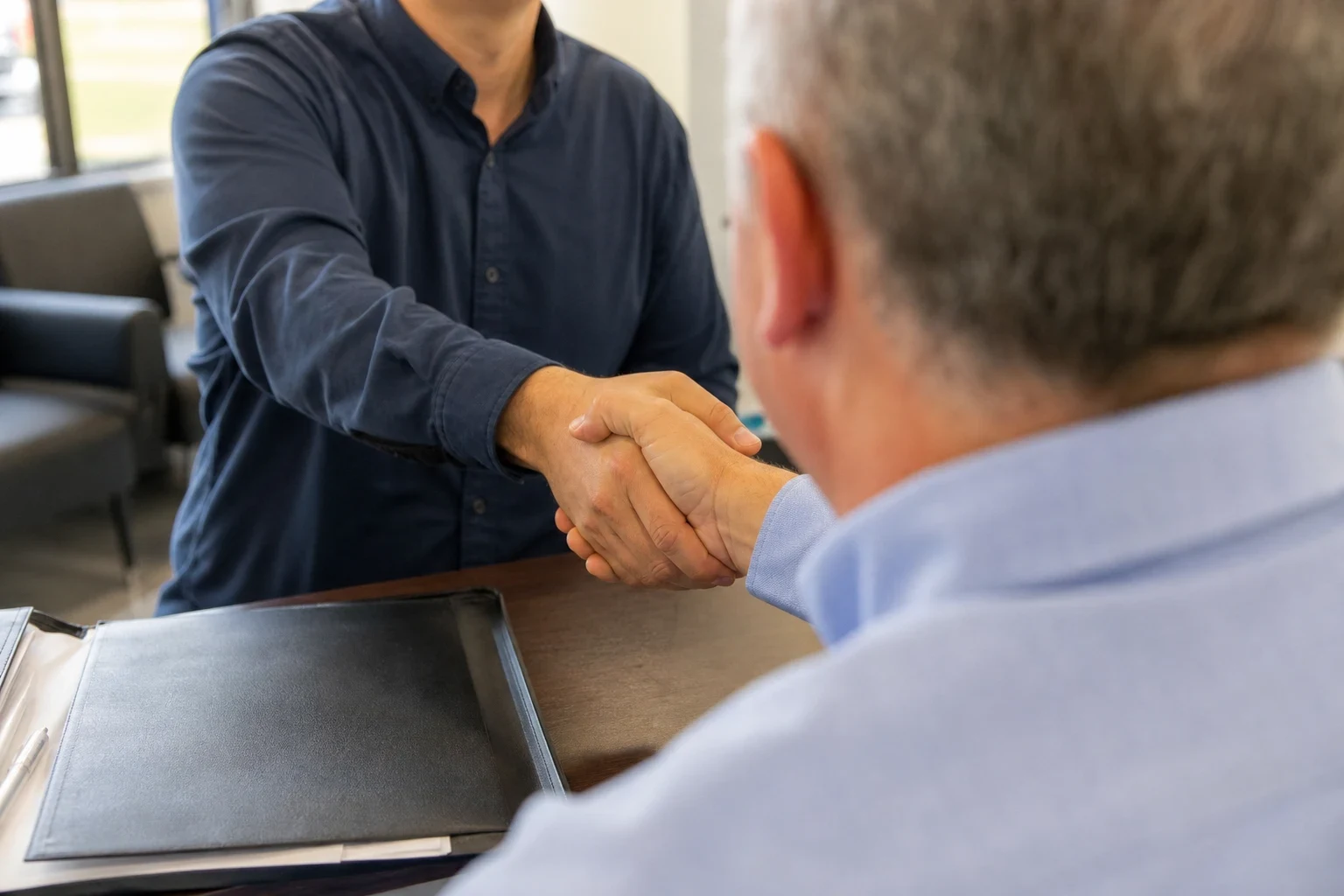 Over-the-shoulder view of a gray-haired client shaking hands with an agent in a navy shirt across a desk with a black leather folder, waiting chairs behind
