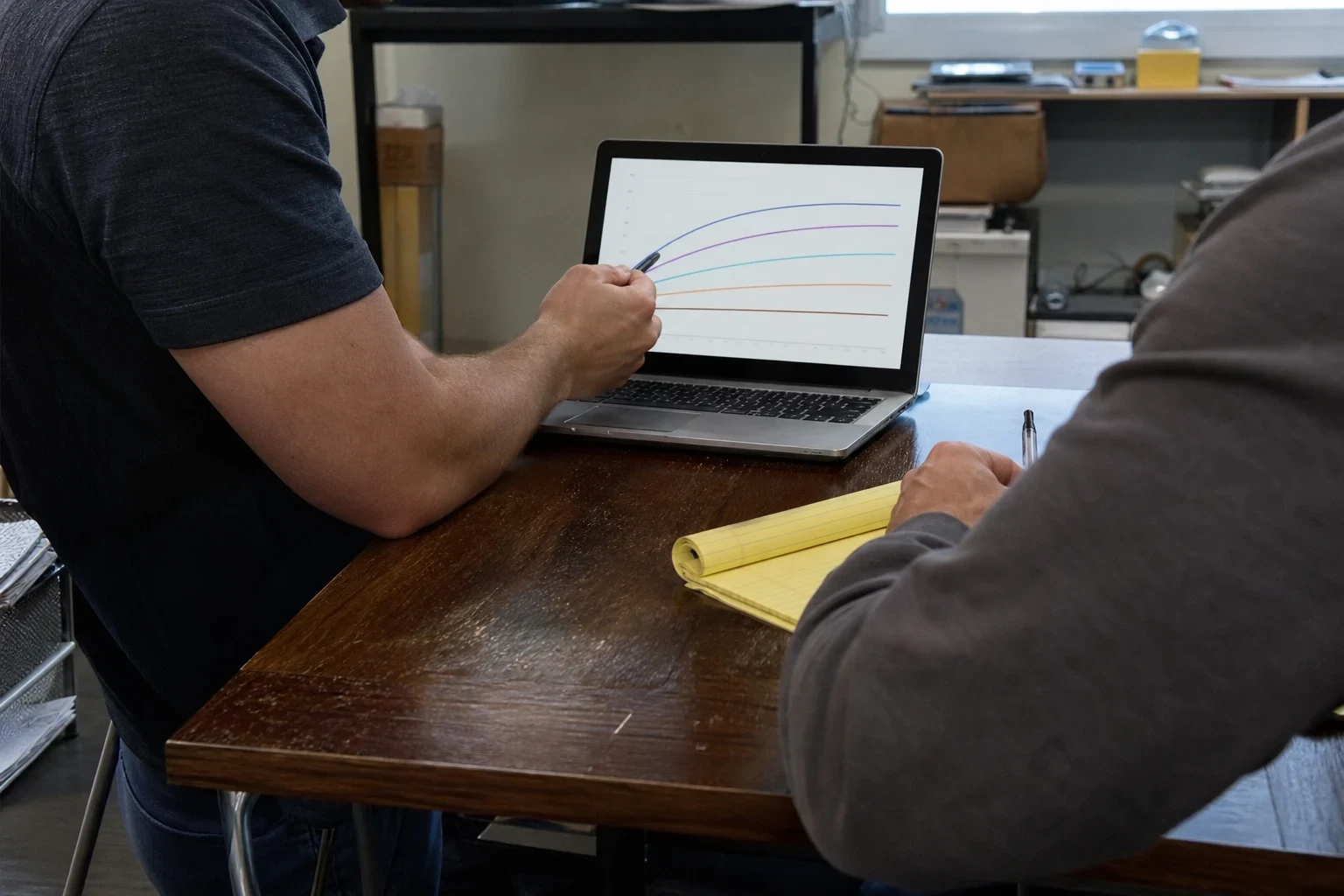 Man in a dark polo points a pen at a laptop graph with curved lines while a client in a gray sweatshirt holds a pen over a yellow legal pad at a worn wooden desk