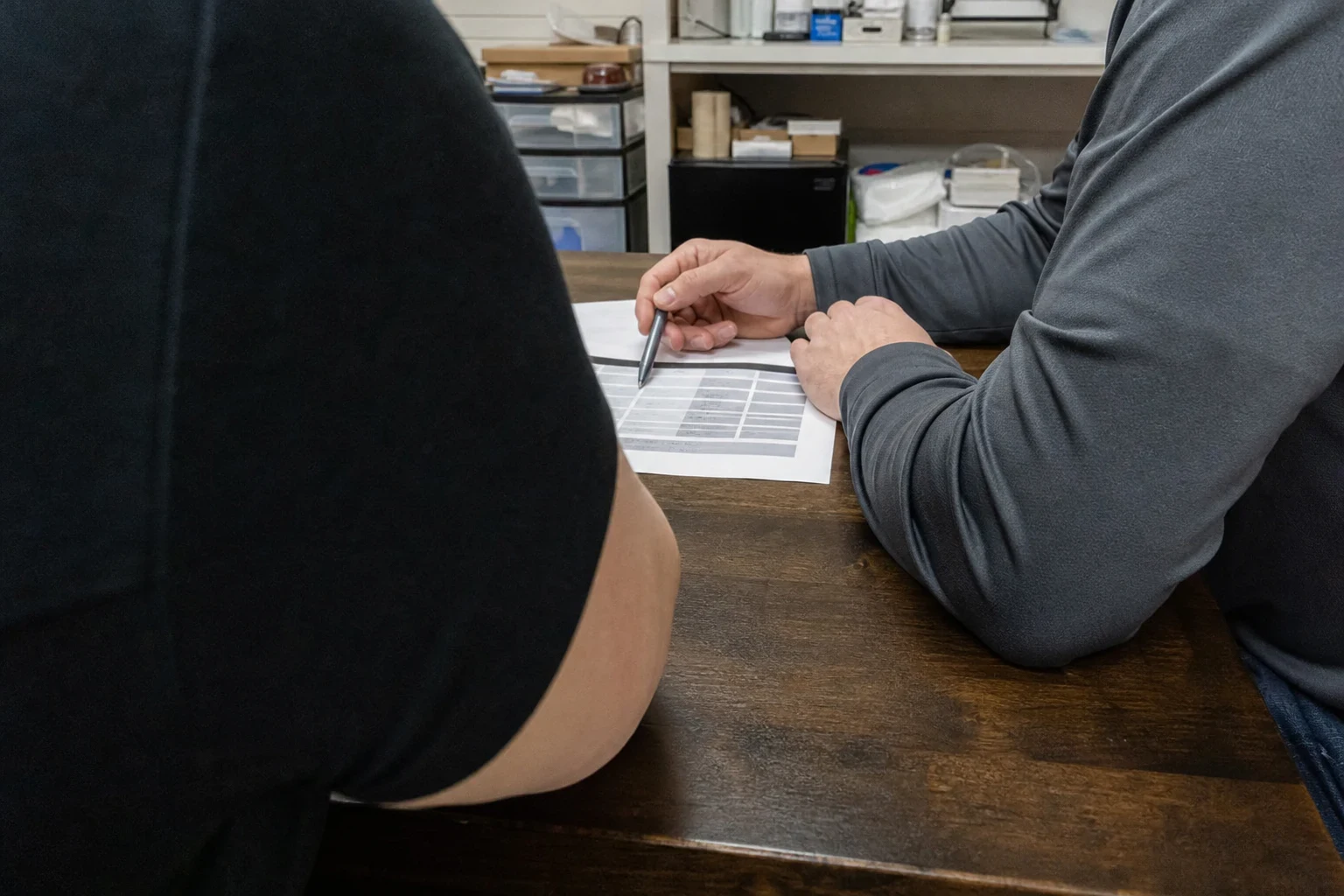 Man in a gray long-sleeve shirt points a pen at a printed table of figures on a wooden desk while a client looks on, with storage shelves in the back office behind